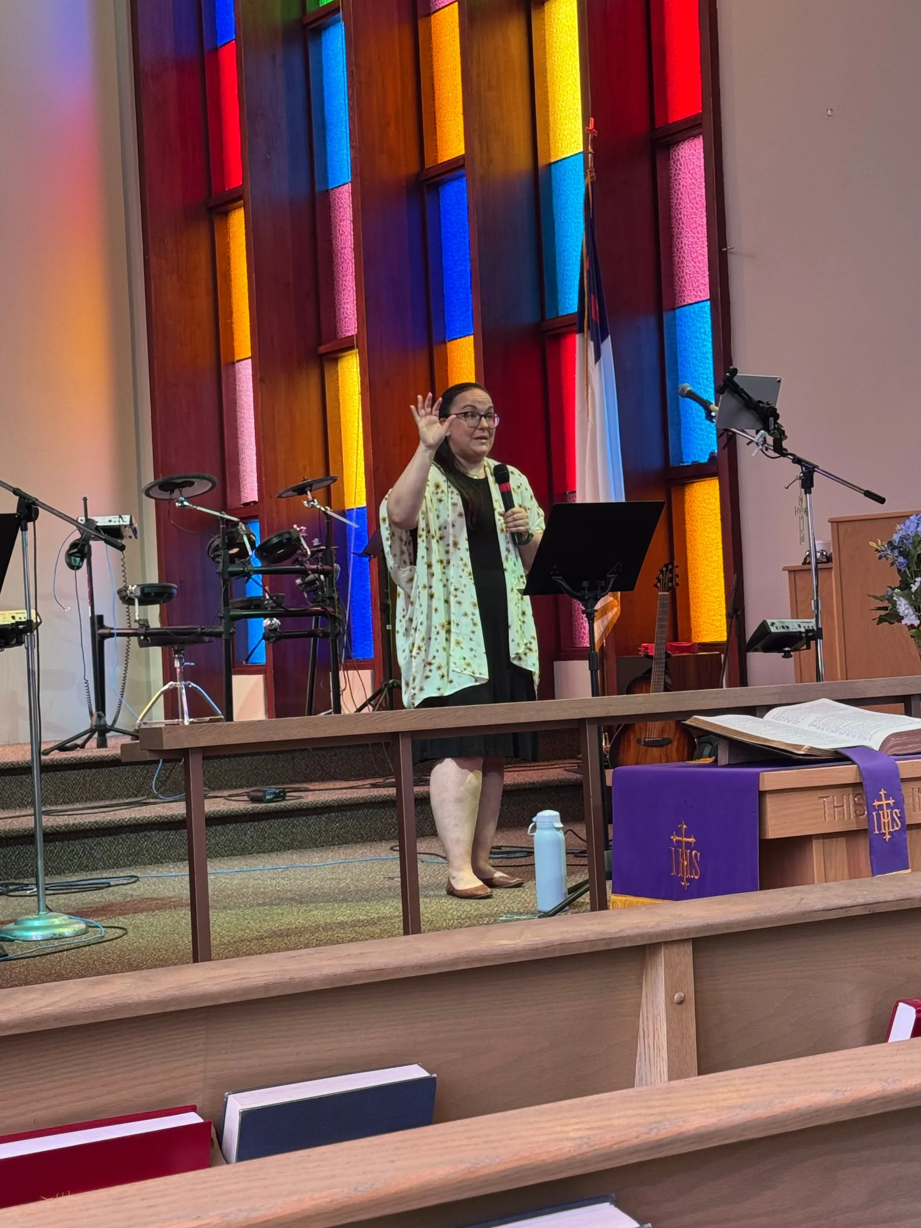 Pastor Ariel speaking with a microphone in TFMC’s sanctuary in front of the stained-glass windows.