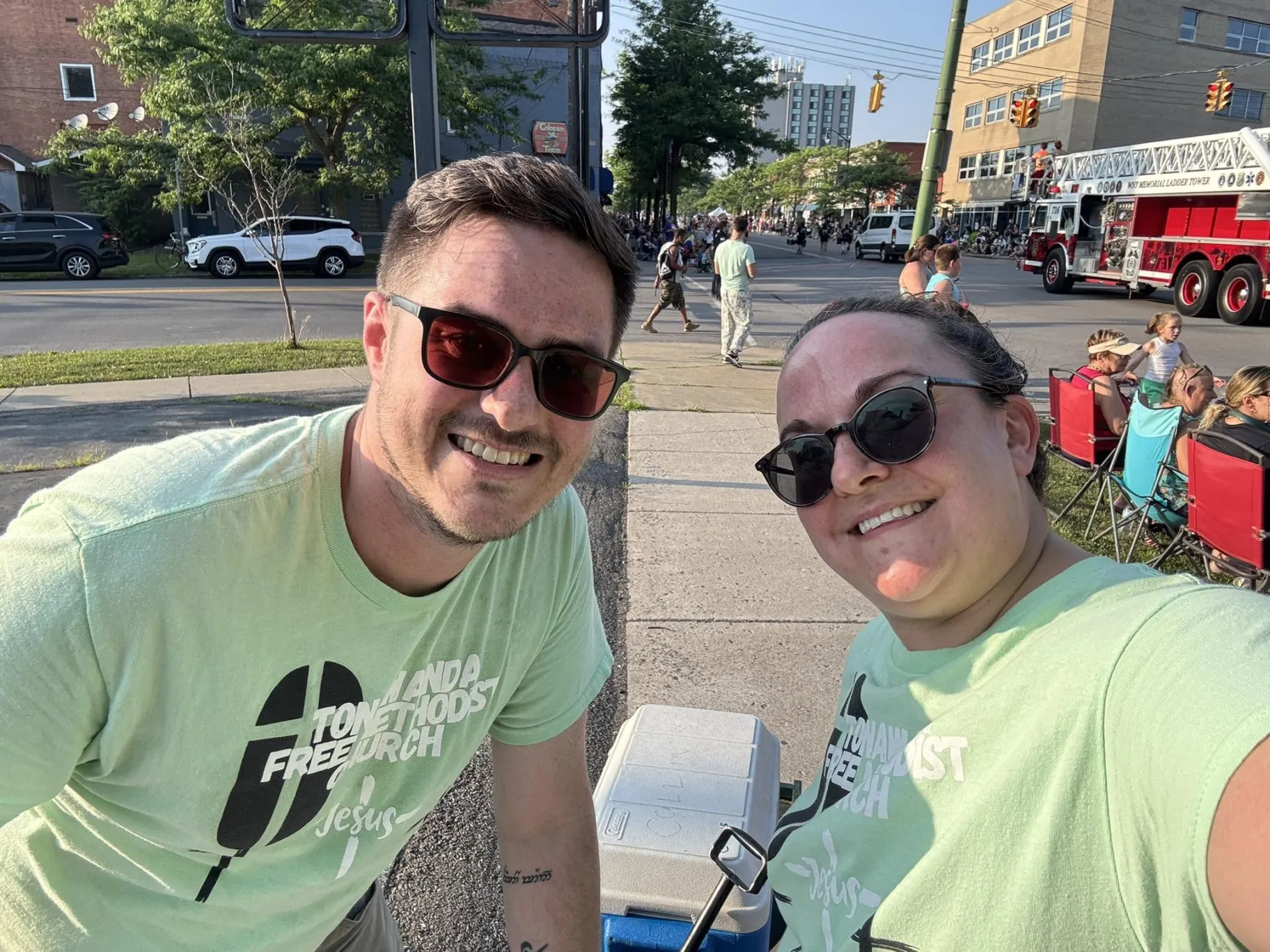 Two smiling TFMC volunteers wearing church shirts at a Tonawanda community parade.